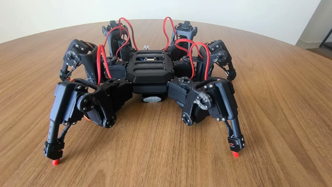 Black 3D-printed hexapod with red braided cables standing on a wooden table.